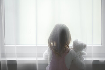 Little girl holding white teddy bear standing alone at window behind transparent day curtains and looking out from home. Rear view. Waiting concept. Close up. - obrazy, fototapety, plakaty