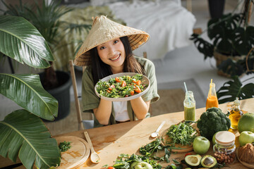 Joyful asian woman in traditional conical hat holding salad from organic vegetables and fruits...