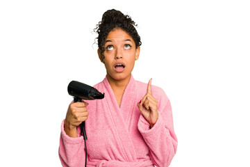 Young brazilian woman wearing a pink bathrobe holding an hairdryer isolated pointing upside with opened mouth.