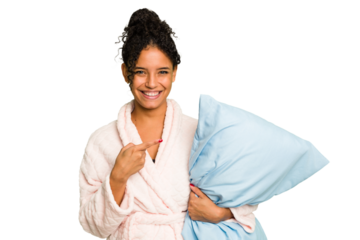 Young brazilian woman wearing a pajama holding a pillow isolated smiling and pointing aside, showing something at blank space.