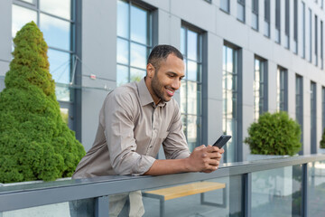 hispanic man outside modern office building using smartphone, businessman in shirt typing message and browsing online pages.