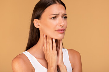 Unhappy caucasian lady suffering from pain in throat, touching her neck, standing over beige background, empty space