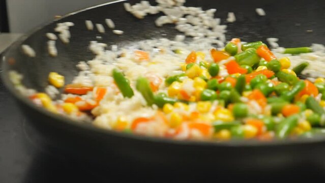 Cloes up of male cook holding frying pan and sauteing a rice with mixed vegetables at kitchen. Chef cooking risotto with corn, peas, asparagus, pepper and carrot on skillet. Concept of preparing food