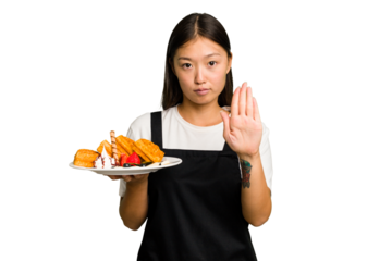 Young asian waitress woman holding waffles isolated standing with outstretched hand showing stop sign, preventing you.