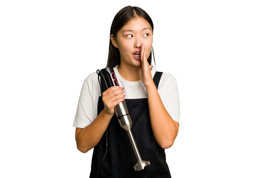 Young asian cook woman holding a blender isolated is saying a secret hot braking news and looking aside