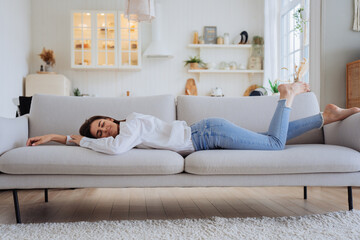 Sleepy woman in white shirt, blue jeans have sleeping on sofa. Tired female lying at home alone in kitchen, feels mental or physical exhaustion. Lack of energy after sleepless night, fatigue concept.