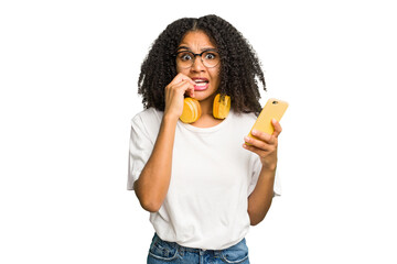 Young african american woman listening to music with yellow headphones isolated biting fingernails,...