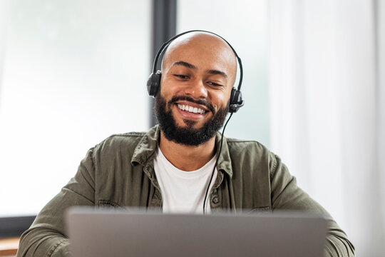 Happy latin man with beard in headphones looking at laptop and smiling, having video call from home