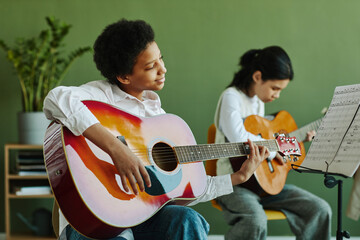 Fototapeta Diligent African American schoolgirl with acoustic guitar looking at paper with notes on music stand while sitting against his classmate obraz