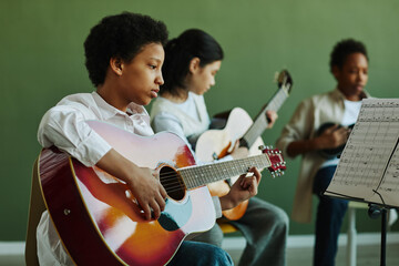 African American schoolgirl with acoustic guitar looking at paper with musical notes while playing...