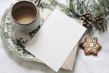 Christmas still life. Blank greeting card, invitation mockup.Gingerbread cookie, cup of coffee and pine tree branches. Floral green plate. White linen tablecloth with envelope. Winter festive flatlay. - obrazy, fototapety, plakaty