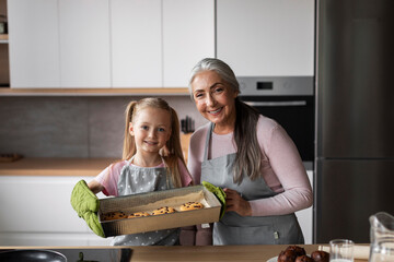 Cheerful caucasian little granddaughter and elderly grandmother show fresh cookies on baking sheet