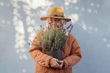 Happy elderly female with potted lavender. Cheerful senior woman in trendy outerwear and hat smiling and carrying pot with blooming lavender on sunny spring day on street