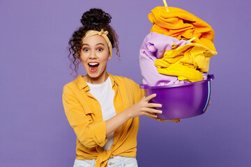 Young shocked happy surprised housekeeper woman wear yellow shirt tidy up hold show basin with clean clothes after laundry isolated on plain pastel light purple background studio. Housework concept. - Powered by Adobe