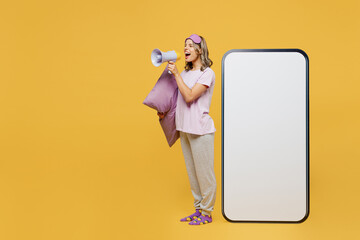 Full body young woman in purple pyjamas jam sleep eye mask rest relax at home big huge blank screen mobile cell phone with area scream megaphone isolated on plain yellow background Night nap concept