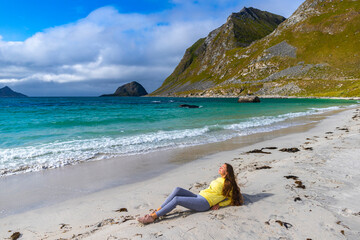 a beautiful long-haired girl sunbathes while sitting on the sand on the famous haukland beach on the lofoten islands in northern norway; relaxing on the beach in the norwegian fjords