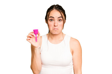 Young caucasian woman holding menstrual cup isolated on green chroma background shrugs shoulders and open eyes confused.