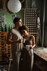 african american woman with cup of tea leaning on dreamy boyfriend looking away in workshop.