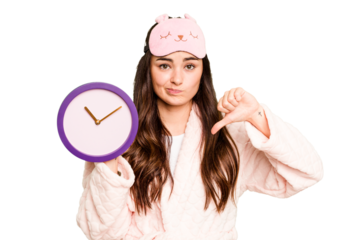 Young caucasian woman holding a clock wearing a pajama isolated on green chroma background showing a dislike gesture, thumbs down. Disagreement concept.
