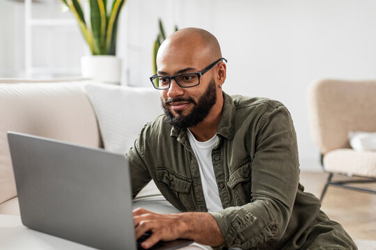 Positive mature latin man using laptop computer, browsing internet or working online, sitting on couch at home