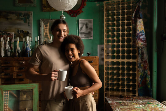 happy interracial couple with tea cups looking at camera in studio with vintage decor.