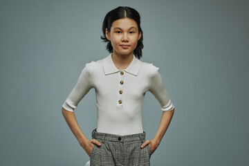 Youthful schoolgirl of Asian ethnicity wearing white tanktop and grey pants keeping hands in pockets and looking at camera - obrazy, fototapety, plakaty