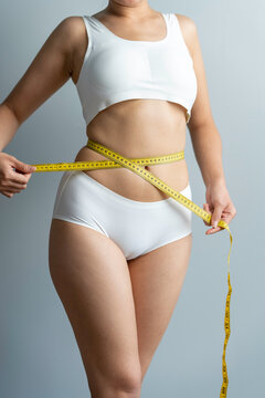 A woman wearing a bra is using waist tape to measure her waist size.