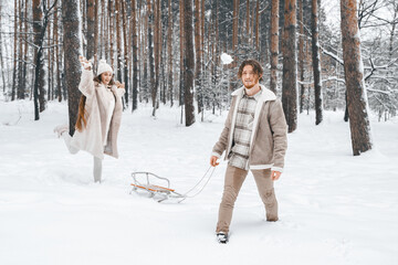 Fototapeta na wymiar Love romantic young couple girl,guy in snowy winter forest sledding,playing snowballs.Walking with sleigh in stylish clothes, fur coat,jacket, woolen shawl,bonnet.Snow lovestory.Romantic date,weekend