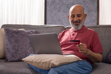 Senior man shopping online through laptop using credit card while sitting on sofa at home
