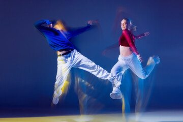 Portrait of young man and woman dancing hip-hop isolated over dark blue background with mixed lights. Jumping high