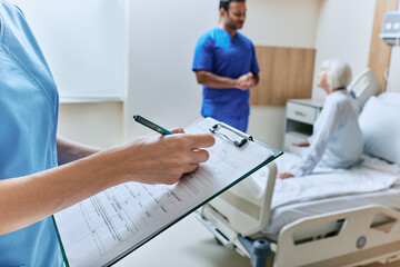 Nurse takes notes in medical chart in hospital room with elderly patient and doctor while bedside consultation. Medical caring about patient