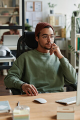 Young modern IT engineer sitting by workplace in front of computer and looking at screen while...