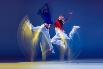 Portrait of young man and woman dancing isolated over dark blue background with mixed lights. Jump...