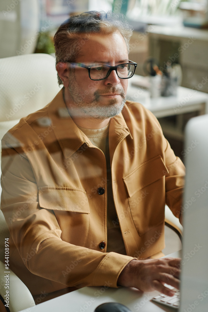 Poster Modern mature webdesigner or programmer sitting by desks in front of computer monitor and looking at screen while working online