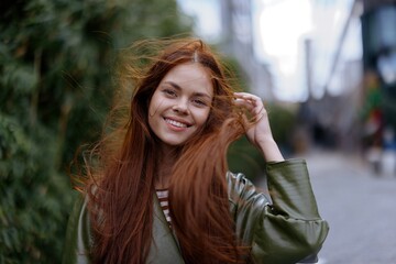 Portrait of a beautiful young woman in the city looking into the camera smile with teeth with red flying hair in fashionable clothes in the city against of bamboo in spring, lifestyle in the city