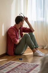 Frustrated European man sits near with mobile phone on floor of own home and sadly looks out...