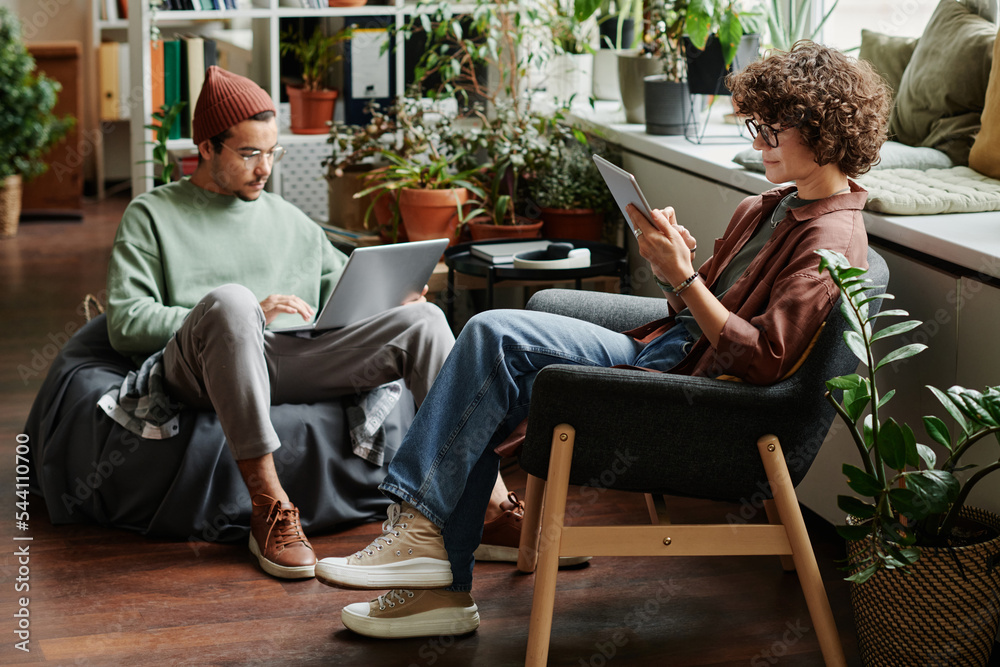 Poster two young serious coworkers using wireless mobile gadgets while sitting in armchairs in openspace of