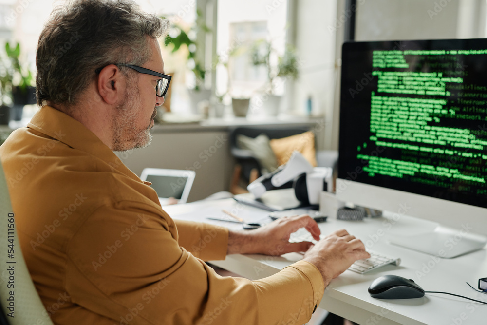 Poster Side view of mature serious male IT engineer typing on computer keyboard while sitting in front of screen with coded data