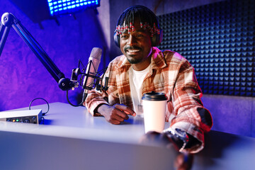 African radio host sitting at desk recording in studio with microphone and laptop