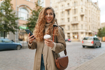 Young woman wearing autumn coat walking with smartphone and coffee cup in a city street