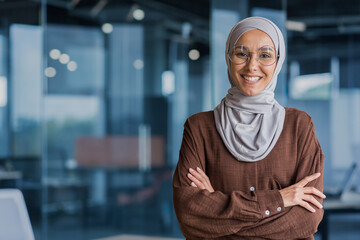 Portrait of successful and happy businesswoman in hijab, office worker smiling and looking at camera with crossed arms, working inside modern office. - obrazy, fototapety, plakaty