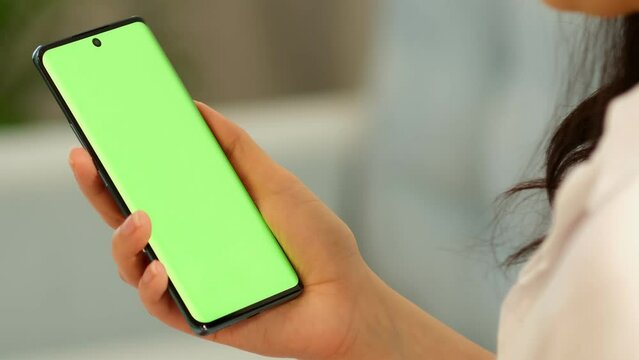 Woman holds mobile phone swipes photos or pictures left indoors of cozy home. Use green screen for copy space closeup. Chroma key mock-up on smartphone in hand.