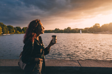 A student girl with a backpack drinks fragrant coffee near the embankment of Lake Aasee in Munster...