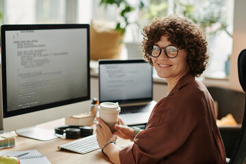 Young successful IT specialist with cup of coffee looking at camera while sitting by workplace with...