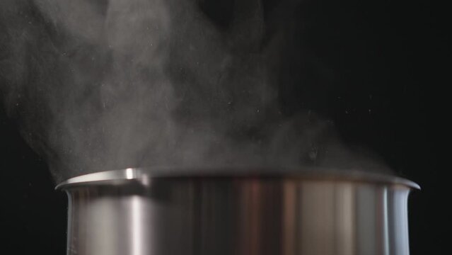 Slow motion closeup of steam rising from steel pot over black background