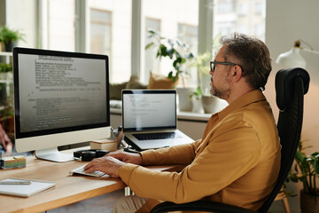 Serious mature software developer looking at code on computer screen while decoding data and...