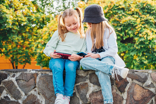 Two smiling little sister girls kids sitting and browsing their smartphone devices in the autumnal park. Careless young childhood time and a modern technology concept image.