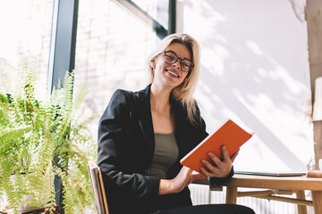 Portrait of cheerful student in smart casual black jacket and optical spectacles smiling at camera during time for learning and studying in cafe, successful woman with literature book posing