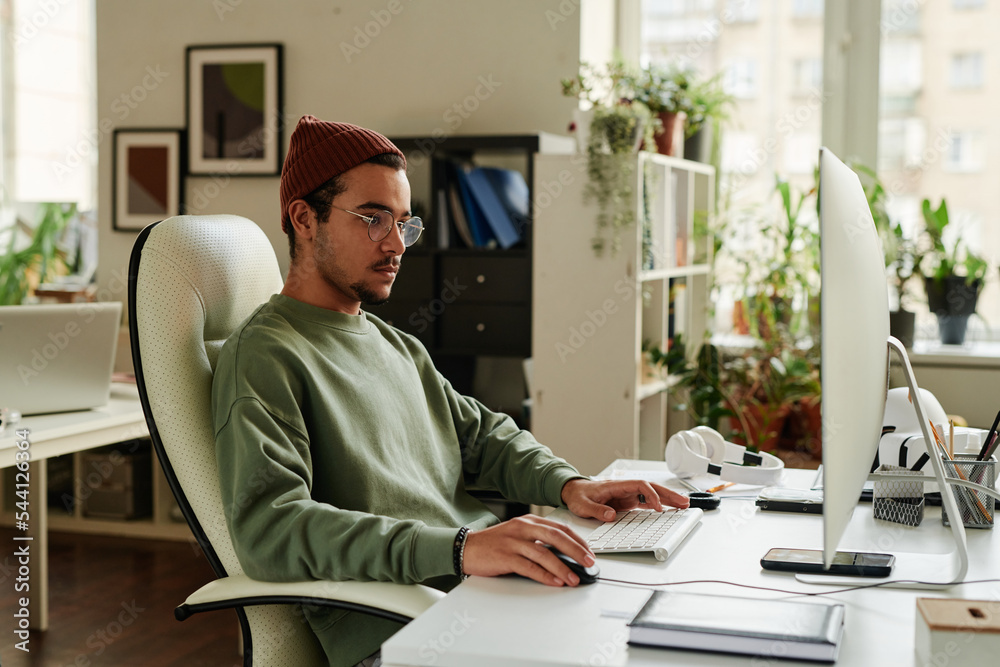 Poster Young serious IT support engineer looking at computer screen and clicking mouse while working over new software by workplace