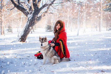 A young woman in a red hooded raincoat with a gray husky dog on the background of a winter forest...